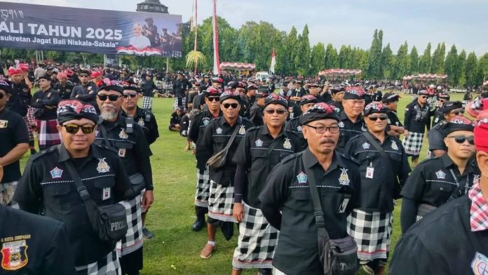 Ratusan Pecalang Bali mengenakan seragam adat hitam dengan kain kotak-kotak dan ikat kepala merah, berdiri di lapangan terbuka pada acara Gelar Agung Pecalang Bali 2025 di Denpasar, dengan bendera merah putih dan spanduk acara di latar belakang.